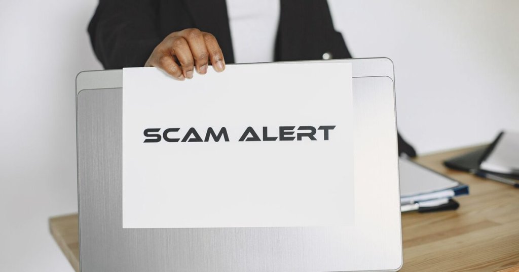A person holding a sheet of paper reading Scam Alert in front of a laptop